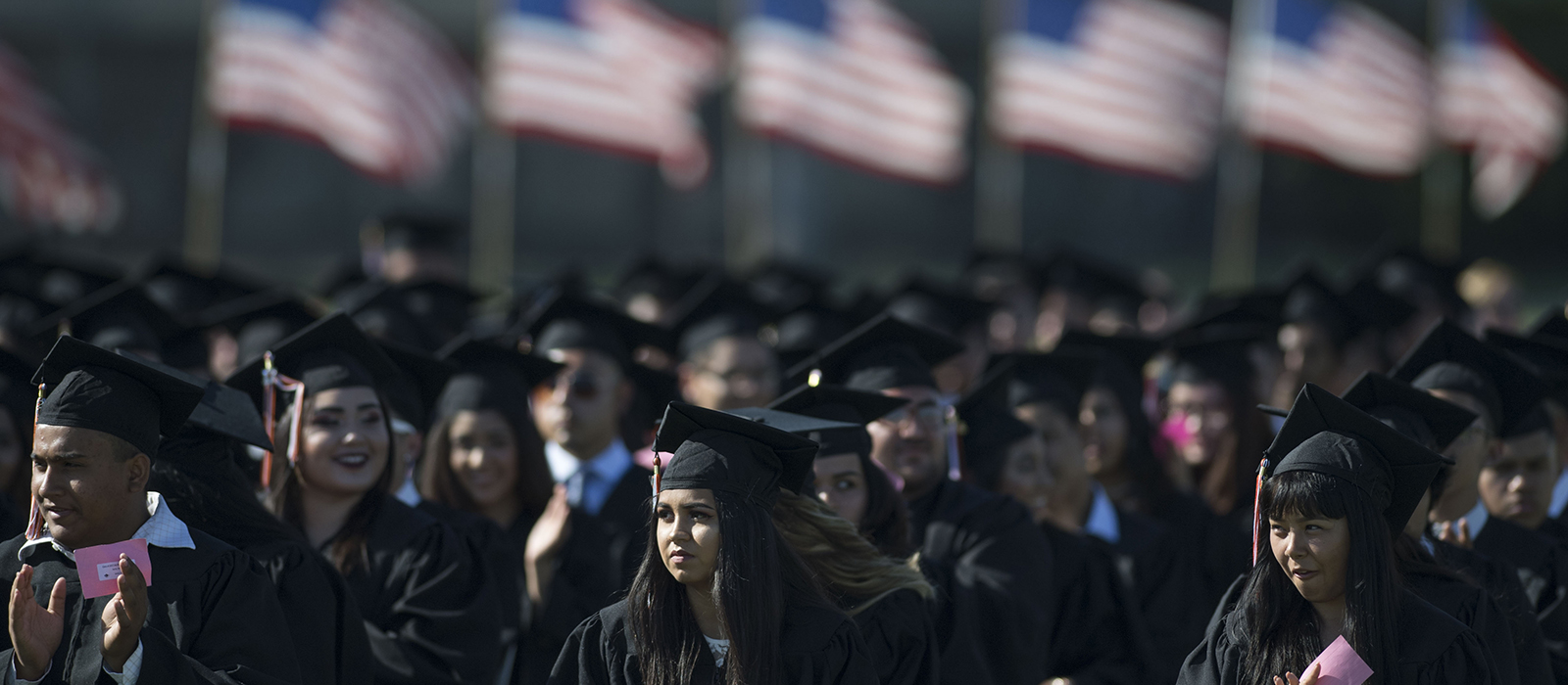 Graduates applaud during a high school graduation, with a row of American flags in the background.