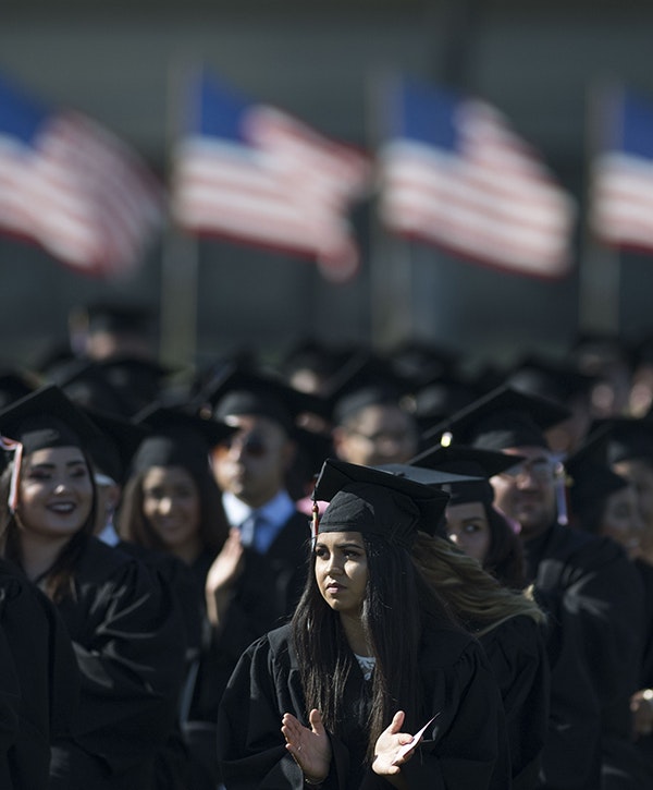 Graduates applaud during a high school graduation, with a row of American flags in the background.