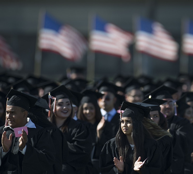 Graduates applaud during a high school graduation, with a row of American flags in the background.