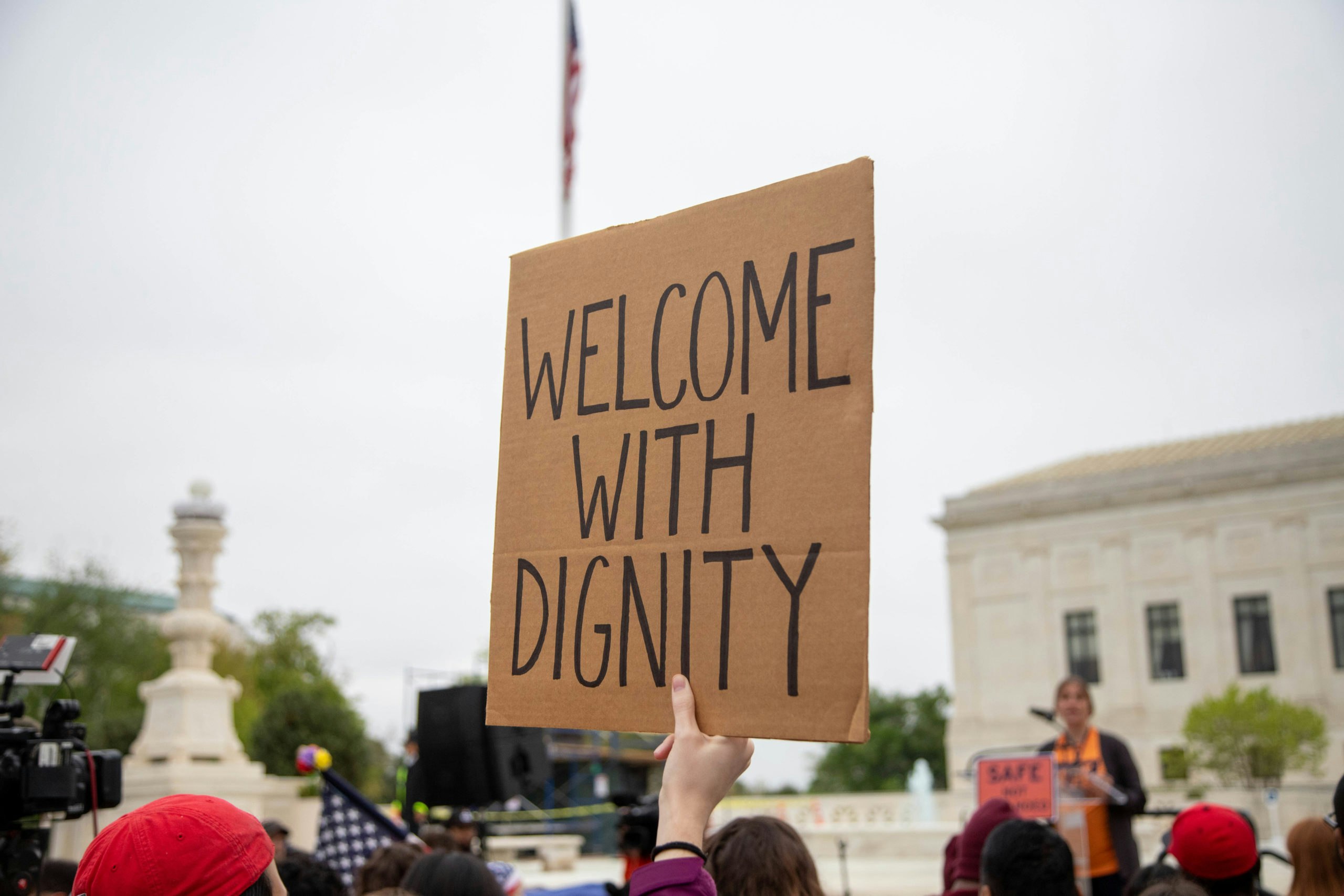 A "Welcome with Dignity" cardboard sign is being held up at the Safe Not Stranded rally at the Supreme Court