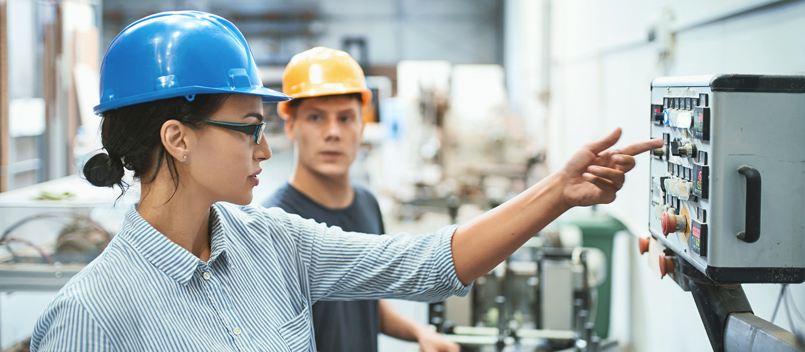 Closeup front view of male and female early 30's engineers at a factory plant standing next to a control panel of the production machine. the woman is explaining the inexperienced worker how to operate it.