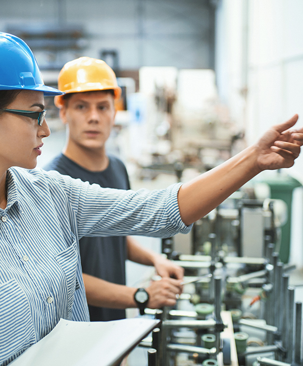 Closeup front view of male and female early 30's engineers at a factory plant standing next to a control panel of the production machine. the woman is explaining the inexperienced worker how to operate it.