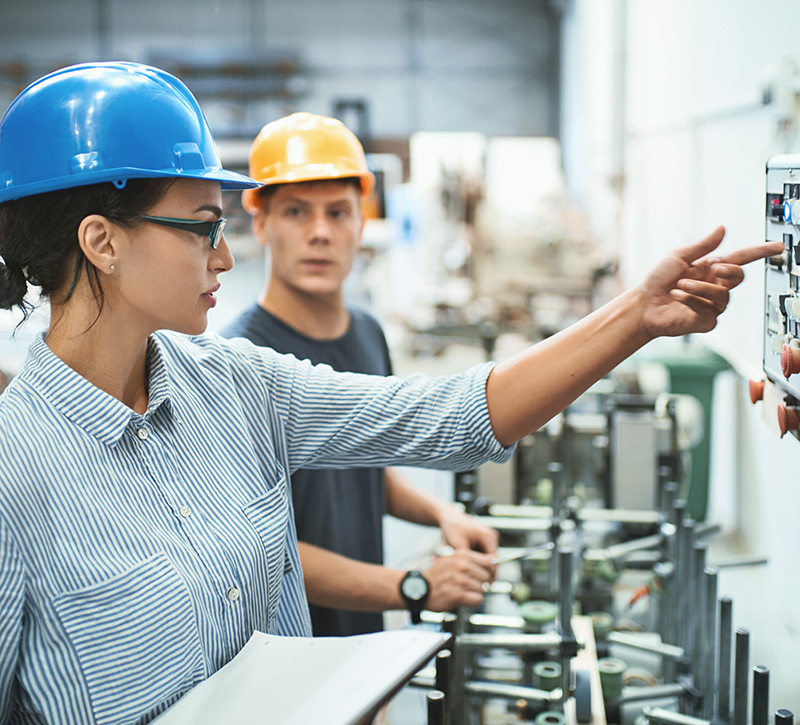 Closeup front view of male and female early 30's engineers at a factory plant standing next to a control panel of the production machine. the woman is explaining the inexperienced worker how to operate it.